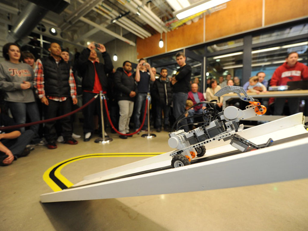 Small, student built LEGO, robot is climbing a ramp in the Introduction to Engineering course competition while spectators watch